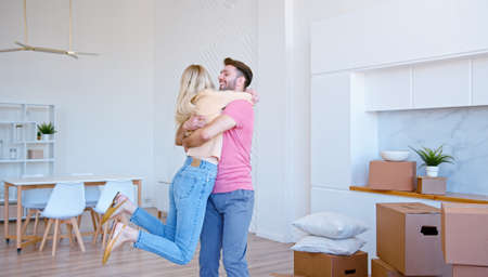 Amazed couple in love puts large cardboard boxes on new apartment floor and hugs rotating with cheerful smiles in empty roomの写真素材
