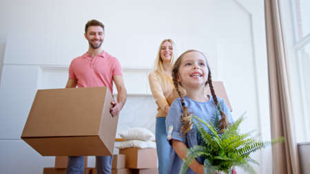 Smiling family man lady and preschooler kid walk along new apartment holding cardboard packages and green pot plant low angle shotの写真素材