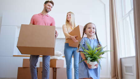 Funny little daughter helps parents with brown cardboard packages move holding small green pot plant in new flat low angle shotの写真素材