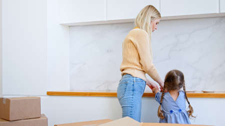 Concentrated preschooler girl with plaits gives blonde mother white plates taken from brown cardboard box in new apartment kitchenの写真素材