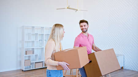 Couple in love looks into eyes and communicates walking along new flat with large boxes in hands against white empty bookshelfの写真素材