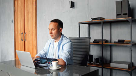 Concentrated man hacker with black bio hand prothesis types on gray laptop sitting at table with cup of coffee in home roomの写真素材