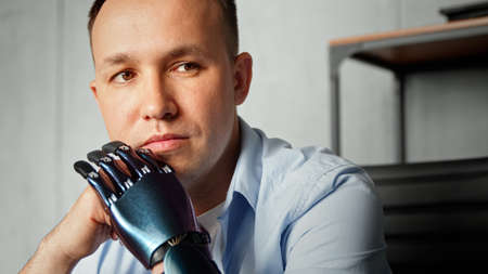Serious cyborg disabled man with bio prothesis thinks holding head on hands sitting against blurred bookshelf at home extreme closeupの写真素材