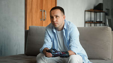 Interested bald guy with artificial hand prothesis switches television channels with remote controller and watches sitting on gray sofaの写真素材