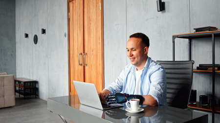 Smiling man programmer with artificial bio hand prothesis types on laptop keyboard sitting at table with cup of coffee at homeの写真素材