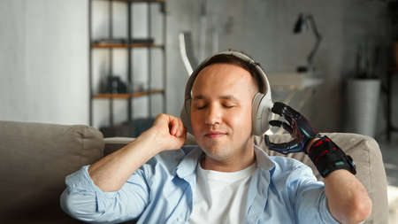 Disabled man with high temple and artificial hand prothesis listens to favorite track in wireless earphones with happy smile at home closeupの写真素材