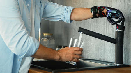 Male person with black artificial hand prothesis pours water into transparent glass from large bowl standing in kitchen at home closeupの写真素材
