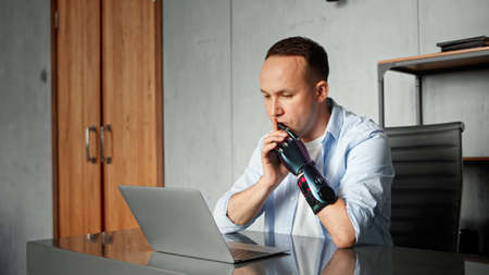 Serious cyborg man programmer with artificial limb prothesis looks around sitting at table with laptop and joining hands at homeの写真素材