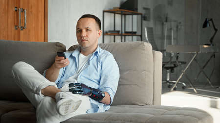Relaxed man with high tech bio hand prothesis takes remote controller and switches television channels sitting on large sofa at homeの写真素材