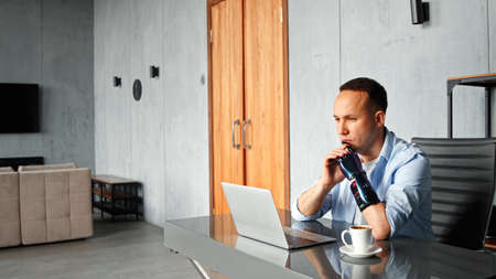 Thoughtful cyborg programmer with black bio hand prothesis looks at laptop screen solving problem sitting at table with cup of coffee in roomの写真素材