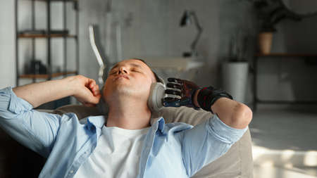 Relaxed disabled man cyborg with black bio hand prothesis listens to music in wireless earphones lying on large sofa at home closeupの写真素材