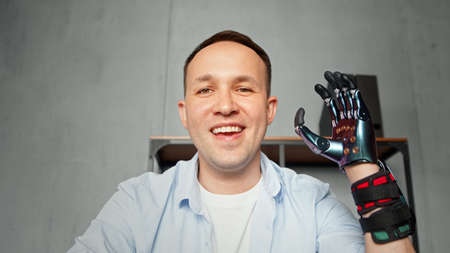 Experienced disabled company worker man waves artificial high tech hand prothesis and smiles at video conference close viewの写真素材