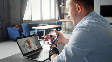 Smiling man with black bio hand prothesis gesticulates talking at online video call with gray haired mother via laptop in roomの写真素材