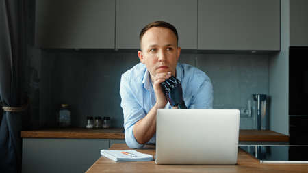 Company manager cyborg guy with artificial hand prothesis sits at table with notes in notebook and laptop solving problem at homeの写真素材