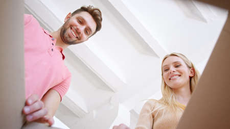 Amazed bearded guy and young woman blonde open brown cardboard box with cheerful smiles and look inside in new flat low angle shotの写真素材