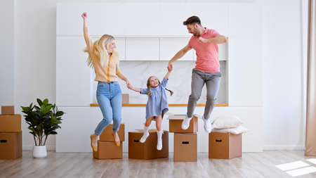 Delighted parents lady and man with preschooler girl jump on new apartment floor joining hands against large cardboard and packagesの写真素材