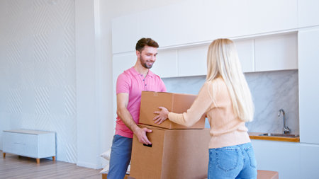 Long haired blonde in pullover puts brown cardboard box in bearded man hands and couple walks from empty new apartment roomの写真素材