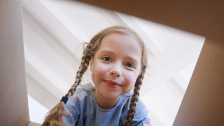 Funny little preschooler girl with long plaits opens brown cardboard box and looks inside after apartment relocation view from boxの写真素材