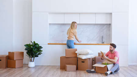 Young woman blonde with long loose hair helps bearded man unpack brown cardboard box in new apartment after relocation to houseの写真素材