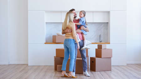 Joyful family with kid girl carries different cardboard boxes into new apartment and puts on large pile on brown wooden floor dancingの写真素材