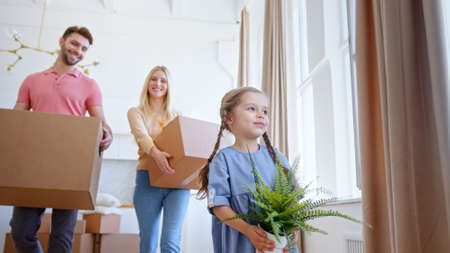 Funny little kid with long plaits carries green pot plant against parents with large cardboard packages walking along new flatの写真素材