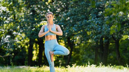 Young woman sportive trainer in blue tracksuit workouts standing in yoga tree pose on green meadow under bright summer sunlightの写真素材