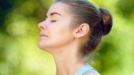 Blonde woman in blue top practices breathing exercises sitting in yoga pose under sunlight in green park super close viewの写真素材