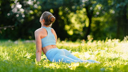 Blonde young woman yoga trainer in blue tracksuit changes upward facing dog pose exercising on meadow under sunlight in summerの写真素材