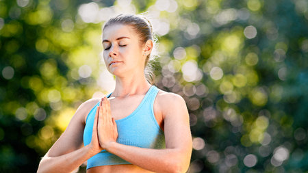 Concentrated lady yoga practitioner in blue top meditates standing in position with closed eyes under bright sunlight in summer closeupの写真素材