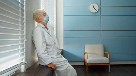 Tired mature female doctor in white robe takes off disposable mask and goggles sitting on windowsill in hospital side viewの写真素材