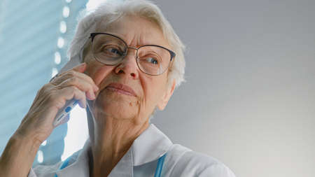 Cheerful senior doctor with eyeglasses talks on mobile phone during distance consultation by window in hospital close viewの写真素材