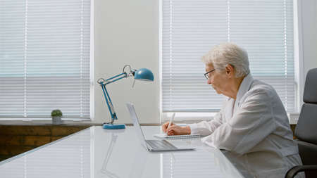 Concentrated mature lady doctor makes notes listening to patient complaints at videochat via laptop in clinic office side viewの写真素材