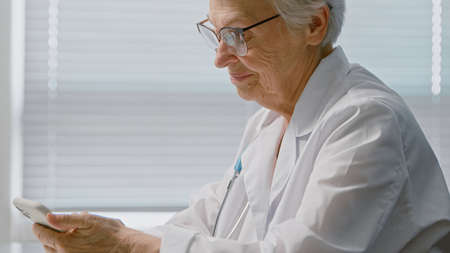 Cheerful senior lady doctor with spectacles texts message with prescription on cellphone sitting near window in clinic closeupの写真素材