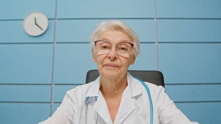 Senior female doctor in robe with elegant glasses consults patient at videocall sitting at table in clinic office camera viewの写真素材