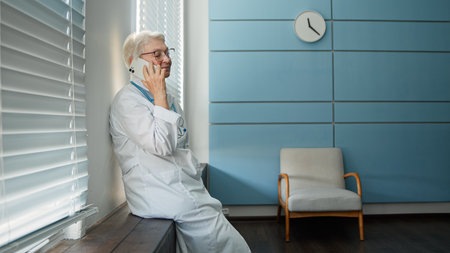 Happy mature lady doctor with glasses talks on cellphone with family member during at short break by window in hospital officeの写真素材