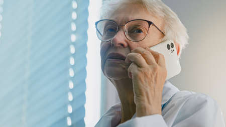 Mature lady doctor with glasses talks on mobile phone giving recommendations to patient in hospital closeup low angle shotの写真素材