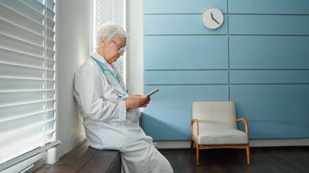 Skilled female doctor in white uniform consults patient at videocall sitting on windowsill in light hospital office side viewの写真素材