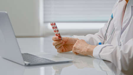 Woman doctor shows blister with small pink pills and explains prescription at videochat via laptop at table in clinic closeupの写真素材