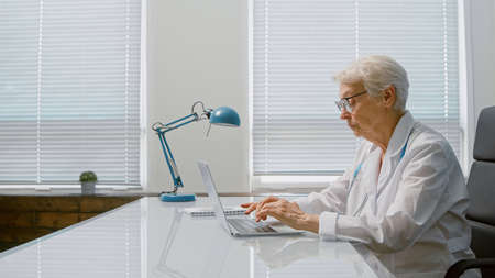 Senior short gray haired lady doctor writes letter on contemporary laptop sitting at table in spacious clinic office side viewの写真素材