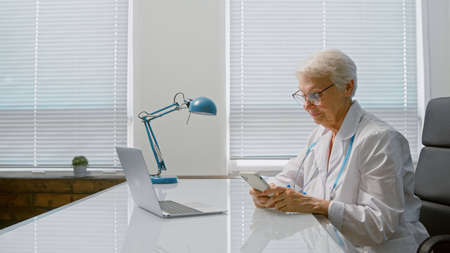 Smiling mature female doctor with spectacles writes message with medical prescription on smartphone at table with laptop in clinicの写真素材