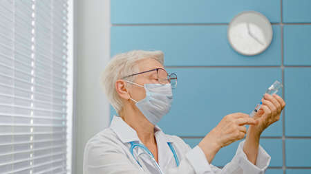 Mature gray haired woman doctor with spectacles and surgical mask takes vaccine from vial with syringe in hospital office closeupの写真素材