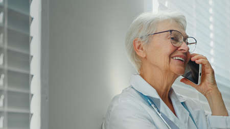 Positive gray haired mature lady doctor with glasses talks on mobile phone with family member near window in hospital roomの写真素材