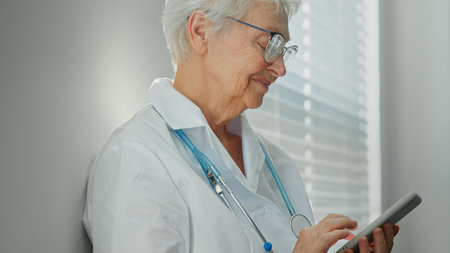 Happy mature female doctor in white robe with stethoscope texts on phone near window in light hospital at break side viewの写真素材