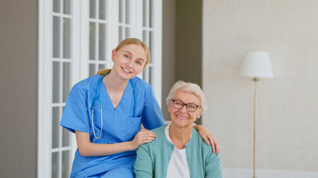 Pretty young doctor with stethoscope hugs smiling senior woman patient posing for camera at contemporary long term care homeの写真素材