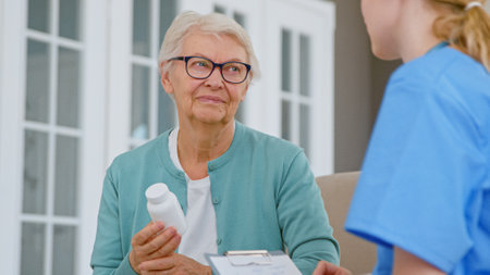 Senior patient woman with glasses holds pills bottle listening to doctor explanations in room at long term care home closeupの写真素材