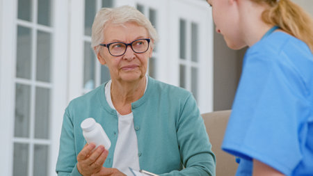 Senior patient woman with glasses holds pills bottle listening to doctor explanations in room at long term care home closeupの写真素材