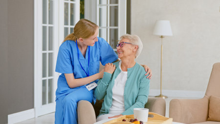Smiling blonde nurse in uniform takes care of senior woman sitting in comfortable armchair at homeの写真素材