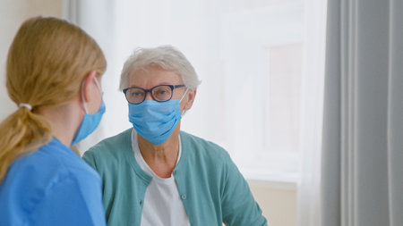 Mature gray haired woman wearing protective mask talks to nurse at appointment with doctor in room at long term care homeの写真素材