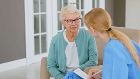 Young blonde doctor interviews short haired senior lady patient making notes in pad at appointment in light clinic office closeupの写真素材