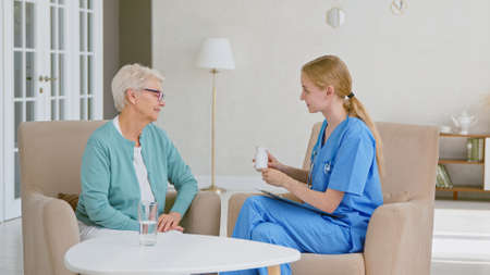 Female doctor shows bottle of medicine to senior patient woman sitting in armchairs in office at modern long term care homeの写真素材